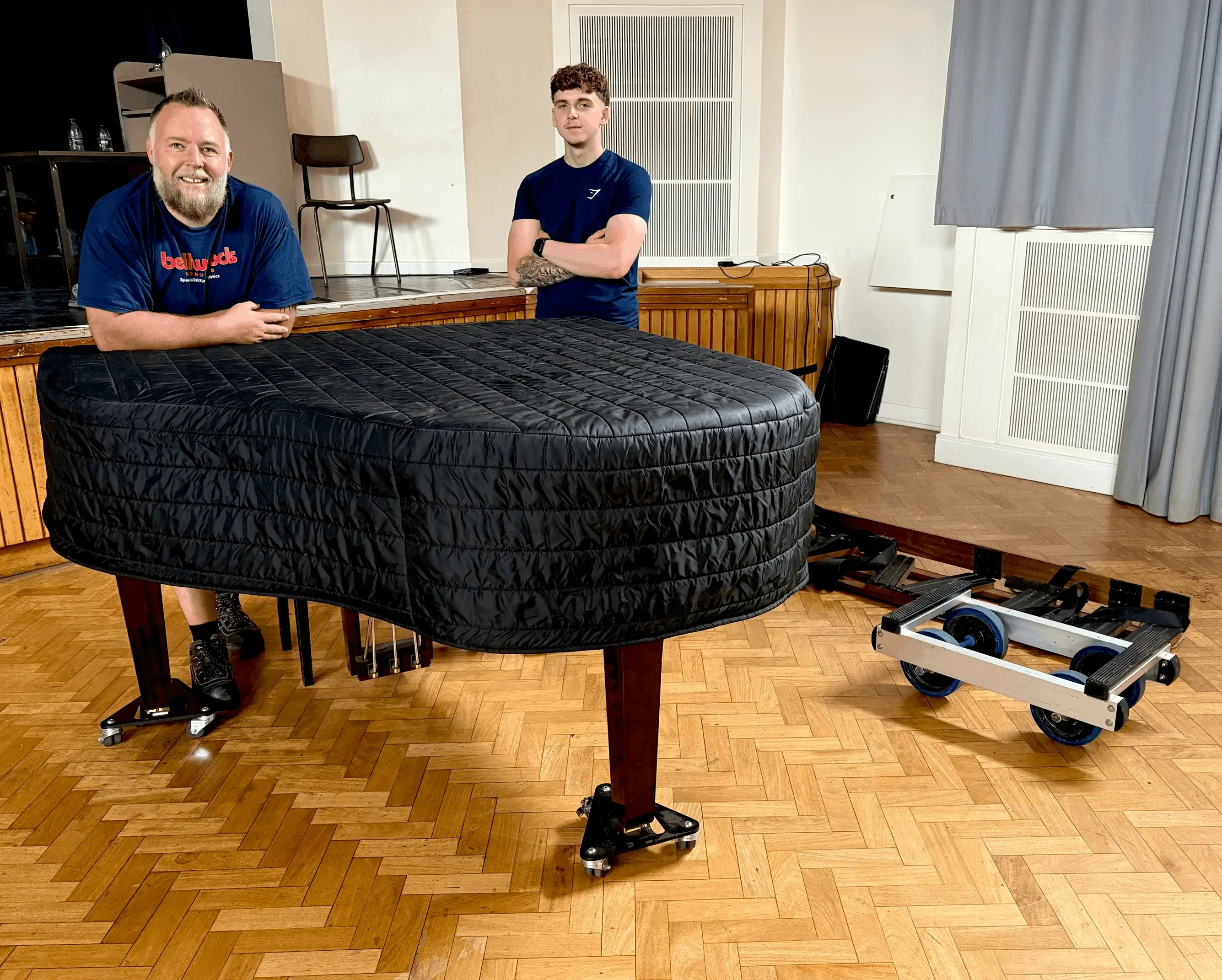 Two Yorkshire Piano Logistics movers beside a grand piano under a protective cover, ready for removal in York