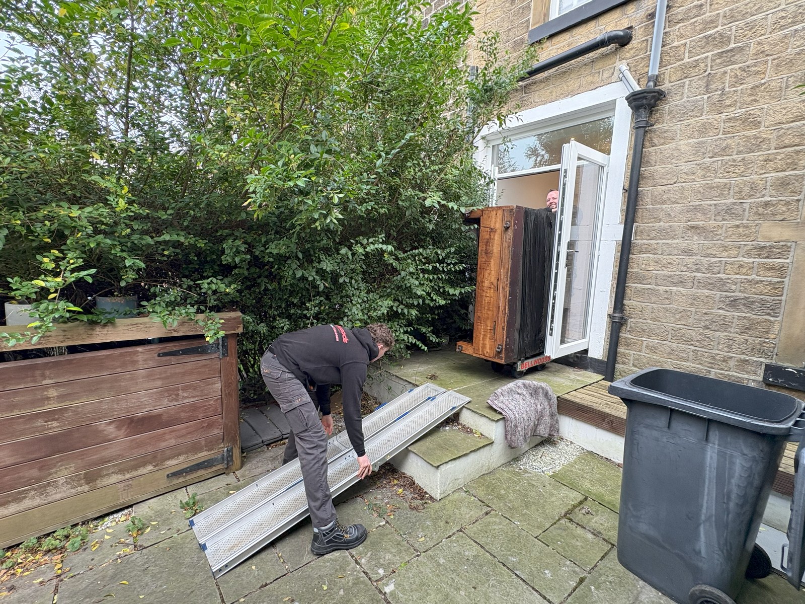 Yorkshire Piano Logistics crew laying an aluminium ramp over garden steps, with a wrapped upright piano waiting on a skate in the doorway